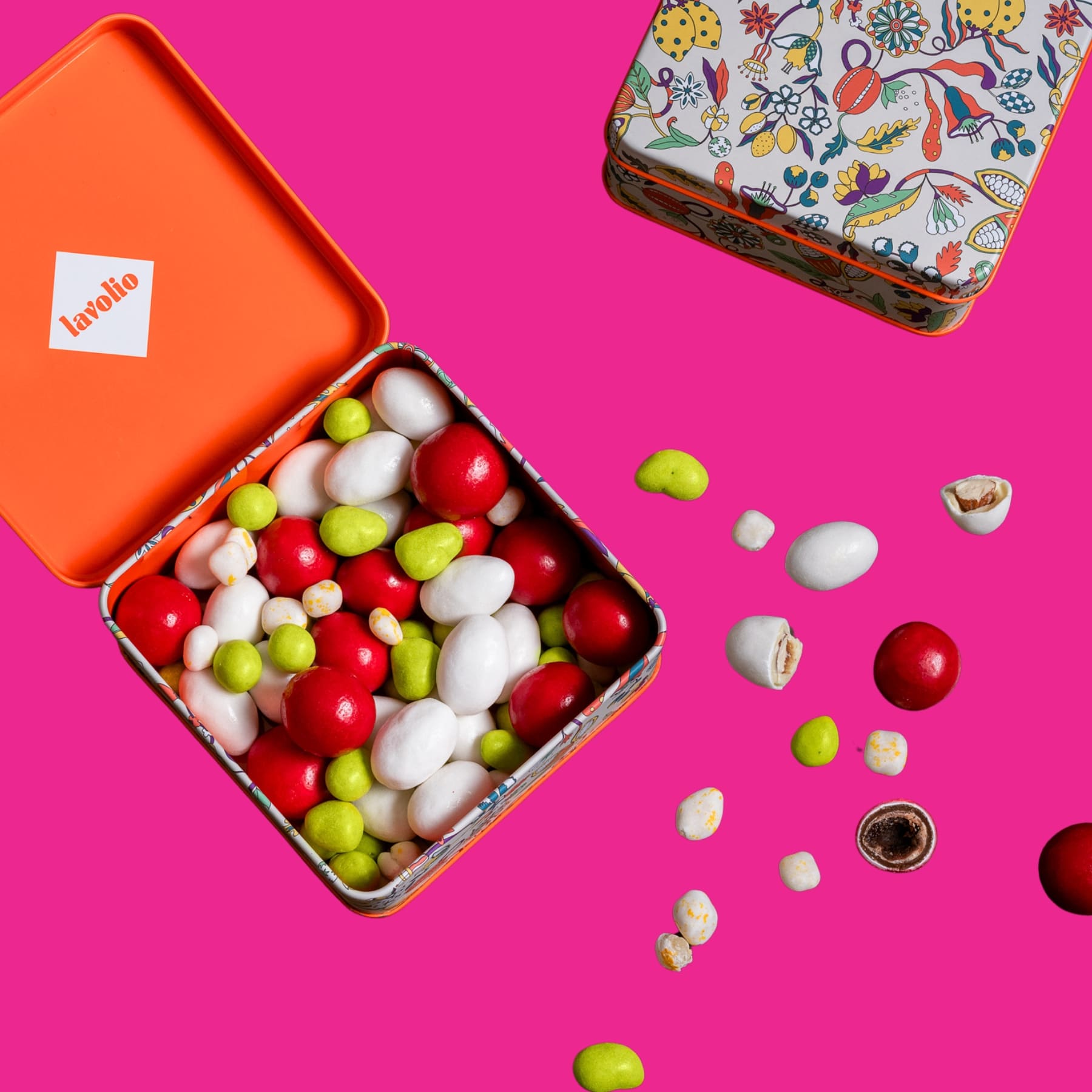 Colorful candy assortment spilling out of an open lavolio orange tin box on a pink background